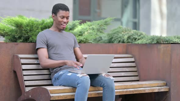 Excited Young African Man Celebrating Success on Laptop on Bench