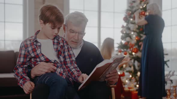 Grandad Reading a Book with Grandson Sitting on a Chair at Christmas Celebration