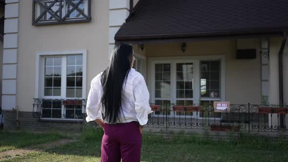 Back View Concentrated Confident Woman with Hands in Pockets Standing in Front of House for Sale