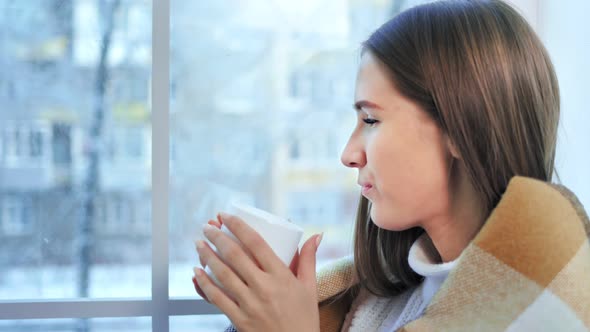 Profile of Young Beautiful Woman Drinking Hot Tea and Looking on Window at Winter Scene