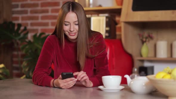 Beautiful Woman Using App on Smartphone and Drinking Coffe in the Kitchen.