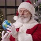 Santa Claus Sits By a Lit Christmas Tree in His Residence Holds a Globe and Turns It Looks - VideoHive Item for Sale