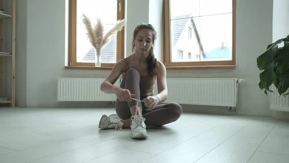 Young Girl in Sportswear Sitting on Floor on Knees Tying Shoelaces on Sneakers