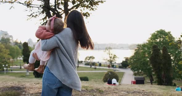 Pretty Young Woman Holding Baby Girl in Her Arms at Sunset at Park. Happy Mother Playing, Hugging