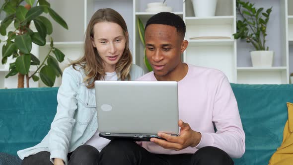 Cheerful Interracial Couple Web Surfing on Laptop Together, Sitting on Sofa at Home