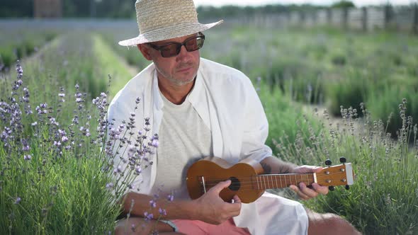 Romantic Caucasian Musician with Ukulele Playing Sitting on Hunkers on Lavender Field Looking Away