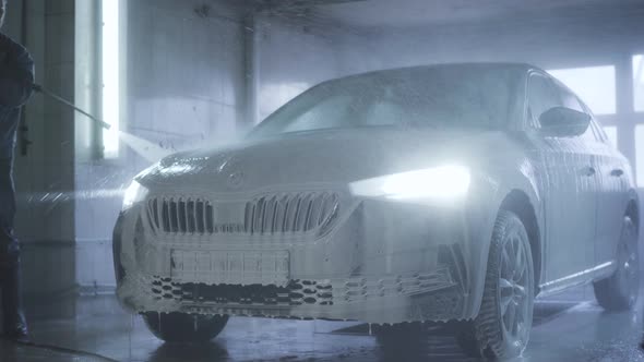 Car with Turned on Headlights Standing in Car Wash Service. Worker Washing Off Foam From Vehicle