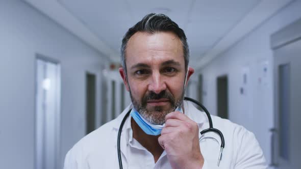 Portrait of smiling caucasian male doctor putting on face mask in hospital corridor