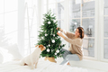 Cheerful woman in comfortable clothes decorates Christmas tree in modern spacious bedroom - PhotoDune Item for Sale
