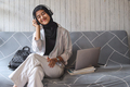 Muslim young woman listening to music with headphones and relaxing while sitting on couch at home. - PhotoDune Item for Sale