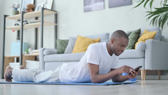 Young African Man Using Smartphone Laying on Yoga Mat