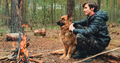 Young man with big dog sits at the fire in the forest. Cheerful guy is stroking his dog, outdoor.  - PhotoDune Item for Sale