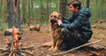 Young man with big dog sits at the fire in the forest. Cheerful guy is stroking his dog, outdoors.  - PhotoDune Item for Sale