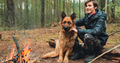 Young man with big dog sits at the fire in the forest. Cheerful guy is stroking his dog, outdoor.  - PhotoDune Item for Sale
