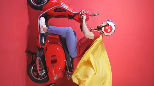 Man in a Helmet and Superhero Costume Rides a Scooter on an Isolated Red Background