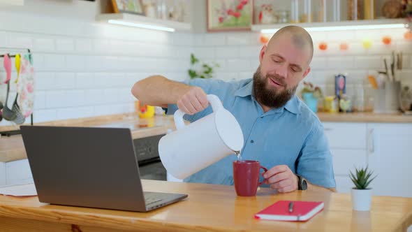 Senior Bearded Man Pours Tea or Coffee Into a Mug While Working or Studying Remotely at Home