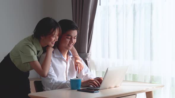 Asian man sitting at the table working on laptop, asian woman walking encourage and help to relax 