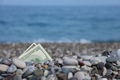 Hundred dollars half covered with round rocks lie on beach - PhotoDune Item for Sale