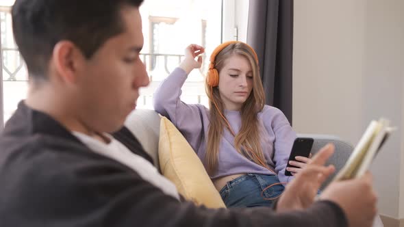 Young couple relaxing and sitting on couch at home while reading a book and listening music