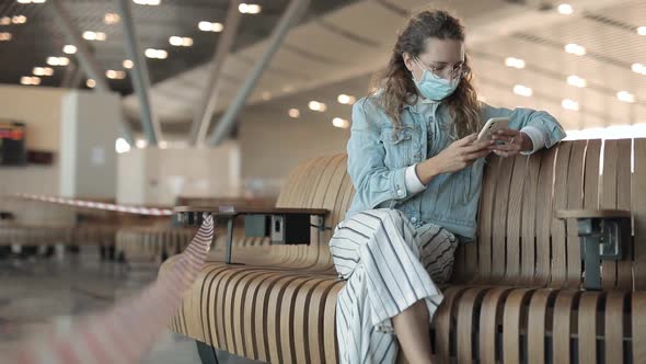 A Young Woman in a Medical Mask Sits with a Phone at the Airport and Waits for Her Departure