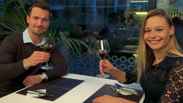 A Man and a Woman Sit in a Restaurant, Clink Glasses, Indicate Clinking with the Camera and Drink