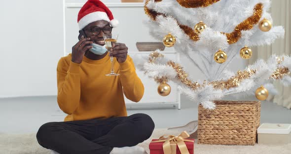 Afro American Guy Black Man Wears Medical Mask Sits Near Christmas Tree in Santa Hat Holding Glass
