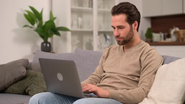 Cheerful Caucasian Man Wearing Casual Wear Sitting on Sofa and Using Laptop at Modern Home Office
