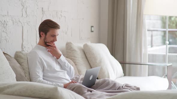 Cheerful Caucasian Man Sits at Home on the Couch and Works Using a Laptop Financial Manager on a