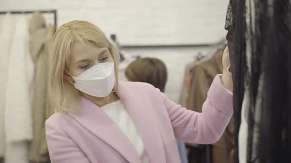 Beautiful Blond Caucasian Woman in Covid Face Mask Choosing Clothes in Shopping Mall and Smiling