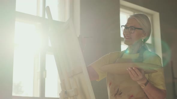 Happy Senior Woman Artist Paints a Picture on Canvas While Standing in a Sunny Workshop