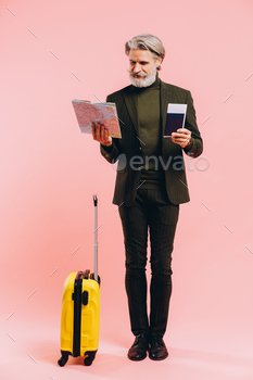 Bearded stylish middle-aged man holding a yellow suitcase, map and passport with a ticket