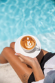Image of Young caucasian woman drinking cappuccino with latte art closed to blue swimming pool - PhotoDune Item for Sale