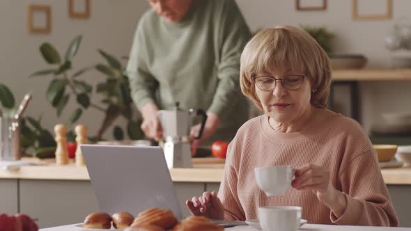 Senior Woman Using Laptop, Drinking Coffee and Talking with Husband
