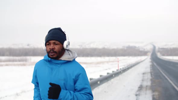 Black Man Wearing Headphones Jogging on Road Next to Highway in Winter Front View