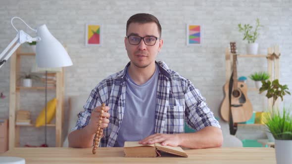 Believing Young Man with a Rosary in His Hands and with a Bible on the Table Looks at the Camera