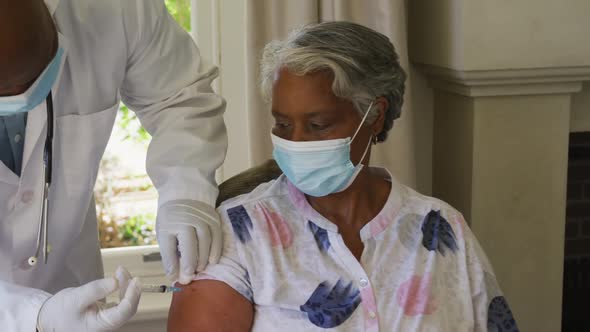 African american senior male doctor injecting covid-19 vaccine into african american woman at home