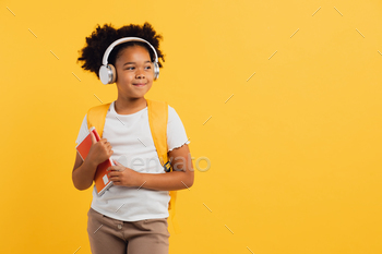 Happy african american schoolgirl in headphones with backpack holding notebooks on yellow background