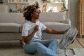 Young African American woman cooling down by ventilator at home, feeling unwell during heatwave - PhotoDune Item for Sale