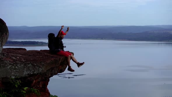 slow-motion of cheerful hiker man sitting and gesture raised arms on the edge of cliff