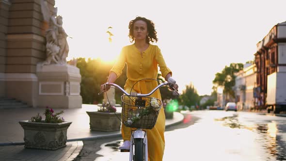 Beautiful Woman Riding a City Bicycle with a Basket and Flowers in the City Center During the Dawn