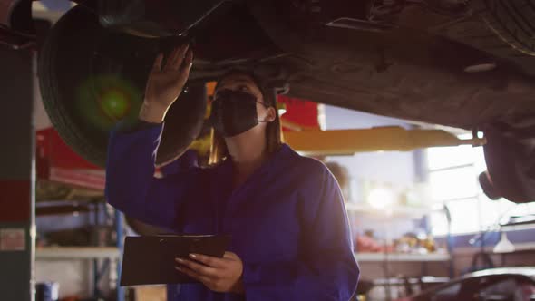 Female mechanic wearing face mask taking notes on clipboard under a car at a car service station