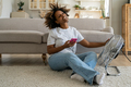 Happy smiling black woman sits on floor with smartphone cools herself with electric ventilator - PhotoDune Item for Sale