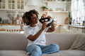 Curious excited young African woman holding modern VR glasses while sitting on sofa in living room - PhotoDune Item for Sale