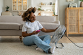 Happy African American woman using smartphone while relaxing in front of electric fan at home - PhotoDune Item for Sale