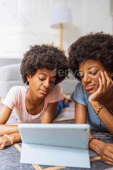 Black twin sisters, using the digital tablet to watch a movie, lying on the living room rug
