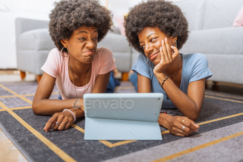 Twin sisters with afro hairstyles, watching boring videos on the digital tablet, lying on the rug