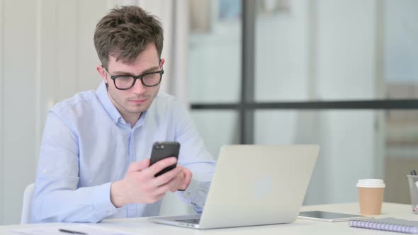 Young Man with Laptop Using Smartphone at Work