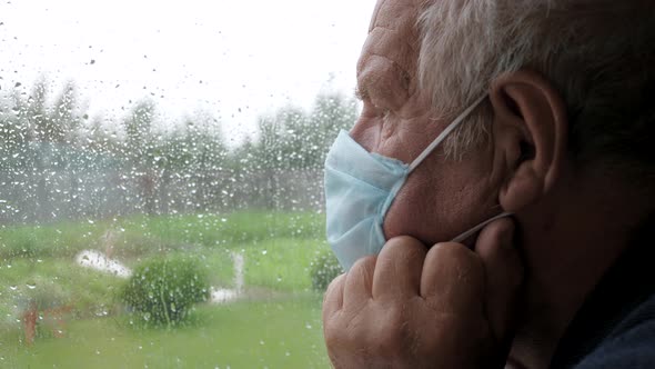 Caucasian Old Sadness Man In Protective Medical Mask Looks Out Window At Rain