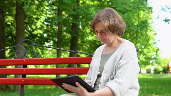 Elderly Woman with a Tablet in Her Hands Sits on a Bench Surfing the Internet and Researching News