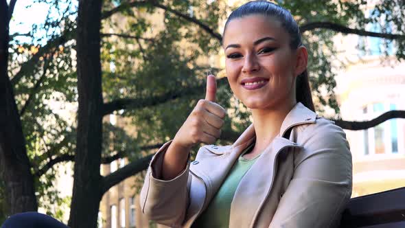 Young Beautiful Woman Sits on the Bench in Park and Shows Thumb Up To Camera